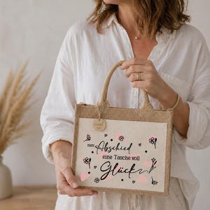 Pu&ograve; includere: Una borsa tote rettangolare in juta con un pannello frontale color crema. Il pannello presenta una scritta nera che recita "zum Abschied eine Tasche voll Gl&uuml;ck" con accenti floreali rosa. La borsa ha un manico e un bordo in juta.