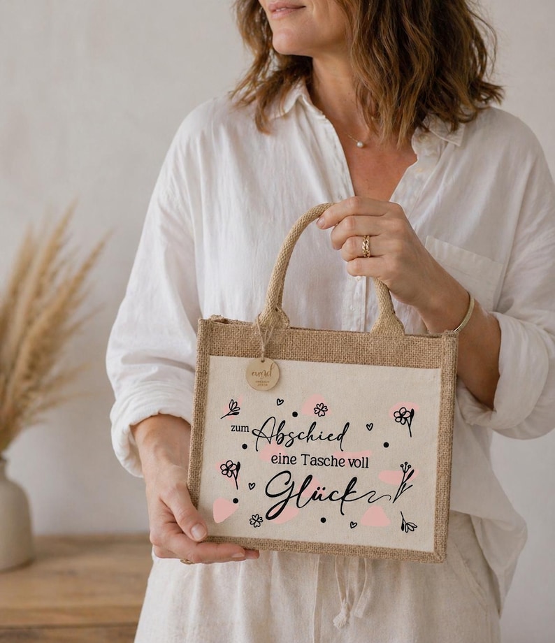 Pu&ograve; includere: Una borsa tote rettangolare in juta con un pannello frontale color crema. Il pannello presenta una scritta nera che recita "zum Abschied eine Tasche voll Gl&uuml;ck" con accenti floreali rosa. La borsa ha un manico e un bordo in juta.