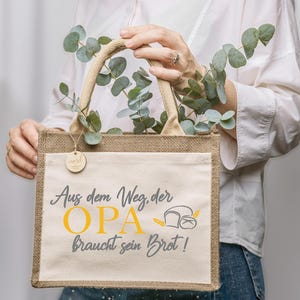 Könnte beinhalten: Eine beigefarbene Jute-Tragetasche mit einer hellbeigen Vordertasche. Die Tasche hat einen Jute-Griff und ein Holzschild. Auf der Vorderseite der Tasche steht der Text "Aus dem Weg, der OPA braucht sein Brot!" in Grau und Gelb.