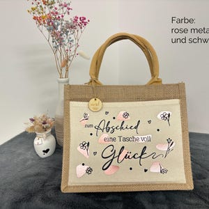 Pu&ograve; includere: Una borsa tote in iuta con un pannello frontale beige chiaro con il testo "zum Abschied eine Tasche voll Gl&uuml;ck" in nero e rosa. La borsa ha un manico marrone e una piccola etichetta di legno.