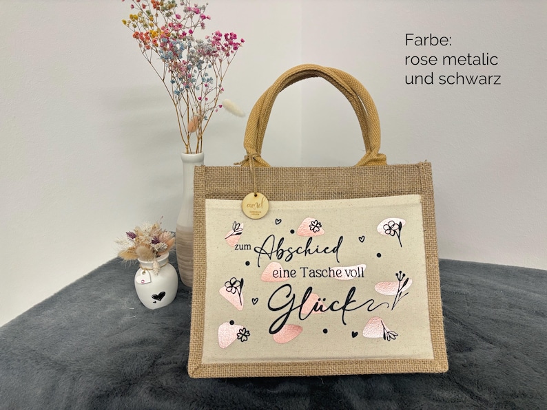 Pu&ograve; includere: Una borsa tote in iuta con un pannello frontale beige chiaro con il testo "zum Abschied eine Tasche voll Gl&uuml;ck" in nero e rosa. La borsa ha un manico marrone e una piccola etichetta di legno.