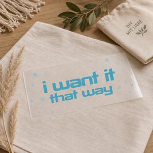 Könnte beinhalten: Ein rechteckiger, transparenter Aufkleber mit dem leuchtend blauen Schriftzug "i want it that way" in einer fetten serifenlosen Schrift, verziert mit kleinen hellblauen Sternen, die über die Oberfläche der Klebefolie verteilt sind.