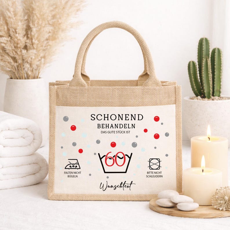 Puede incluir: Un bolso de yute beige con dos asas y un panel frontal blanco. El panel presenta el texto "SCHONEND BEHANDELN DAS GUTE ST&Uuml;CK IST" sobre un "60" rojo grande dentro de un icono de lavabo, con s&iacute;mbolos de lavado y "Wunschtext" debajo.