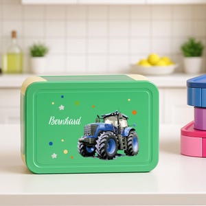 Könnte beinhalten: Eine grüne Brotdose mit einem blauen Traktorgrafik und dem Namen "Bernhard" in weißer Schrift. Die Brotdose hat einen gelben Rand und bunte Stern- und Punktakzente. Weitere Brotdosen sind im Hintergrund gestapelt.