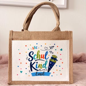 May include: A rectangular jute tote bag with two top handles. The front features a white panel printed with the text "Schul Kind" and "Emil" in blue, surrounded by colourful dots, a paper airplane, hearts, and a blue cone-shaped gift bag.