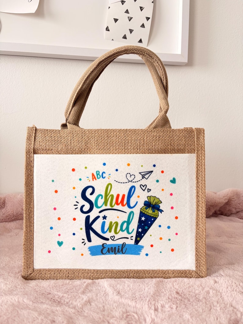 May include: A rectangular jute tote bag with two top handles. The front features a white panel printed with the text "Schul Kind" and "Emil" in blue, surrounded by colourful dots, a paper airplane, hearts, and a blue cone-shaped gift bag.
