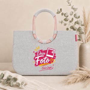 Könnte beinhalten: Eine hellgraue Tragetasche mit zwei ovalen Griffen in metallischem Roségold. Auf der Vorderseite befindet sich eine Grafik mit dem Text "Dein Foto" und "Dein Logo" in Weiß und Pink, ergänzt durch ein kleines Fotorahmen-Symbol.