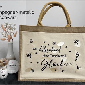 Pu&ograve; includere: Una borsa tote beige con finiture e manici in iuta naturale. La parte anteriore presenta una tasca color crema con la scritta nera che recita "zum Abschied eine Tasche voll Gl&uuml;ck" con accenti floreali e a forma di cuore. Il testo "Farbe champagner-metalic und schwarz" &egrave; in alto a sinistra.