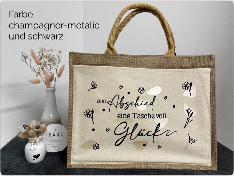 Pu&ograve; includere: Una borsa tote beige con finiture e manici in iuta naturale. La parte anteriore presenta una tasca color crema con la scritta nera che recita "zum Abschied eine Tasche voll Gl&uuml;ck" con accenti floreali e a forma di cuore. Il testo "Farbe champagner-metalic und schwarz" &egrave; in alto a sinistra.
