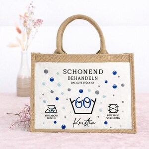 Puede incluir: Un bolso tote rectangular de yute con dos asas y un panel frontal blanco. El panel presenta puntos azules y grises, el texto "SCHONEND BEHANDELN DAS GUTE ST&Uuml;CK IST", un s&iacute;mbolo de lavado a 60 grados, el nombre "Kerstin" e iconos de cuidado textil.