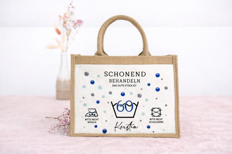 Puede incluir: Un bolso tote rectangular de yute con dos asas y un panel frontal blanco. El panel presenta puntos azules y grises, el texto "SCHONEND BEHANDELN DAS GUTE ST&Uuml;CK IST", un s&iacute;mbolo de lavado a 60 grados, el nombre "Kerstin" e iconos de cuidado textil.