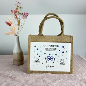 Puede incluir: Un bolso de yute rectangular con dos asas y un panel frontal blanco. El panel presenta el texto "SCHONEND BEHANDELN DAS GUTE ST&Uuml;CK IST 50" con s&iacute;mbolos de lavado, "FALTEN NICHT B&Uuml;GELN", "BITTE NICHT SCHLEUDERN" y el nombre "Andrea".