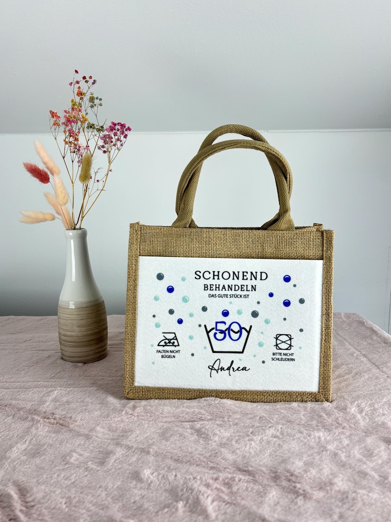 Puede incluir: Un bolso de yute rectangular con dos asas y un panel frontal blanco. El panel presenta el texto "SCHONEND BEHANDELN DAS GUTE ST&Uuml;CK IST 50" con s&iacute;mbolos de lavado, "FALTEN NICHT B&Uuml;GELN", "BITTE NICHT SCHLEUDERN" y el nombre "Andrea".