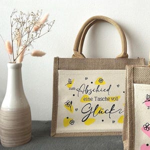Pu&ograve; includere: Una borsa tote in iuta naturale con una tasca frontale color crema. La tasca presenta il testo tedesco "zum Abschied eine Tasche voll Gl&uuml;ck" in scrittura nera e dettagli gialli. La borsa ha un manico e finiture in iuta abbinati.