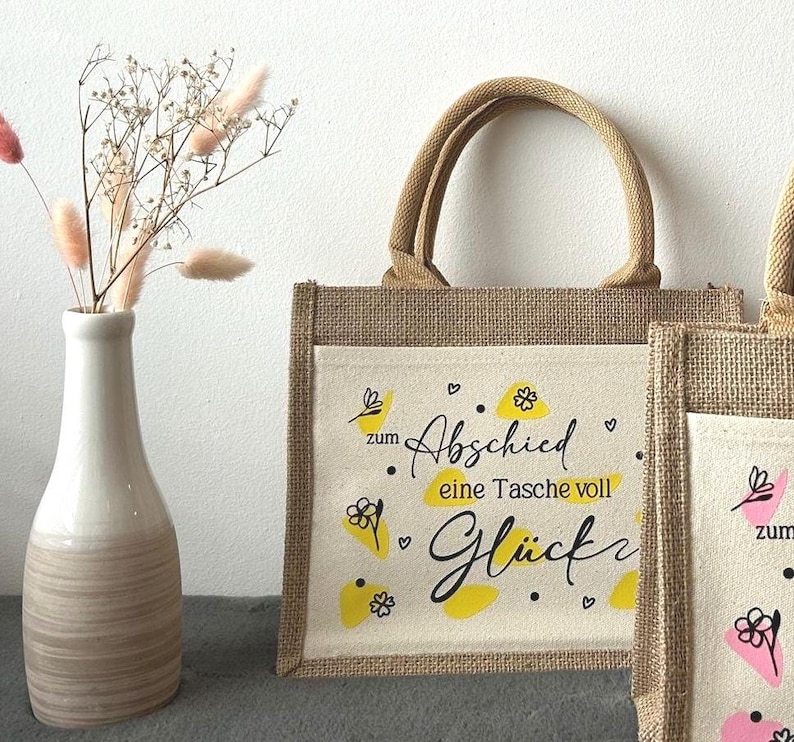 Pu&ograve; includere: Una borsa tote in iuta naturale con una tasca frontale color crema. La tasca presenta il testo tedesco "zum Abschied eine Tasche voll Gl&uuml;ck" in scrittura nera e dettagli gialli. La borsa ha un manico e finiture in iuta abbinati.