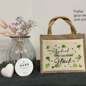 Pu&ograve; includere: Una piccola borsa di juta con una tasca frontale color crema con la scritta "zum Abschied eine Tasche voll Gl&uuml;ck" in nero e dettagli verdi. Sono visibili anche un vaso di vetro trasparente con fiori secchi e un oggetto bianco a forma di cuore.