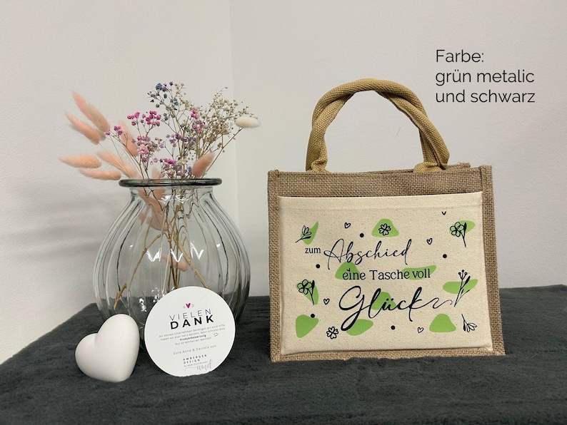 Pu&ograve; includere: Una piccola borsa di juta con una tasca frontale color crema con la scritta "zum Abschied eine Tasche voll Gl&uuml;ck" in nero e dettagli verdi. Sono visibili anche un vaso di vetro trasparente con fiori secchi e un oggetto bianco a forma di cuore.
