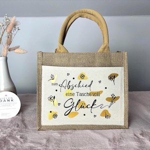 Pu&ograve; includere: Una piccola borsa tote rettangolare in iuta con una tasca frontale color crema. La tasca presenta scritte nere e disegni floreali gialli. La borsa ha un manico in iuta abbinato.