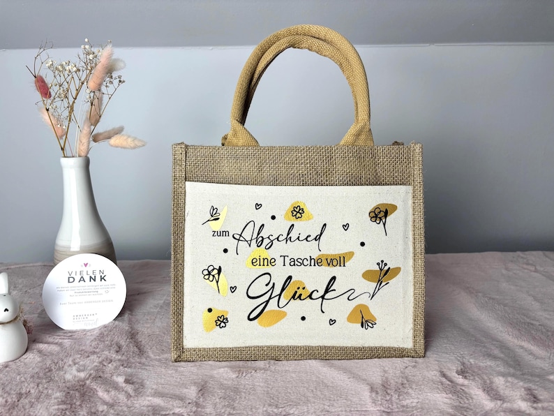 Pu&ograve; includere: Una piccola borsa tote rettangolare in iuta con una tasca frontale color crema. La tasca presenta scritte nere e disegni floreali gialli. La borsa ha un manico in iuta abbinato.