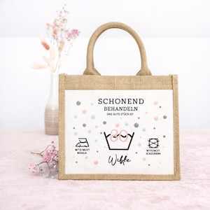 Puede incluir: Un bolso tote rectangular de yute con dos asas y un panel frontal blanco. El panel presenta el texto SCHONEND BEHANDELN, DAS GUTE ST&Uuml;CK IST, el nombre Wibke y s&iacute;mbolos de cuidado de lavado, incluyendo un icono de 30 grados, no planchar y no centrifugar.