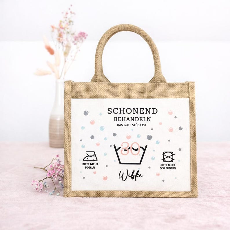 Puede incluir: Un bolso tote rectangular de yute con dos asas y un panel frontal blanco. El panel presenta el texto SCHONEND BEHANDELN, DAS GUTE ST&Uuml;CK IST, el nombre Wibke y s&iacute;mbolos de cuidado de lavado, incluyendo un icono de 30 grados, no planchar y no centrifugar.