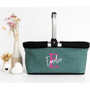 Könnte beinhalten: Türkisfarbener Stoffkorb mit schwarzem Besatz und silberfarbenem Griff. Der Korb trägt den Namen "Emelie" in weißer Schrift mit einem rosa "E". Dekorative Vasen und Blumen sind im Hintergrund.