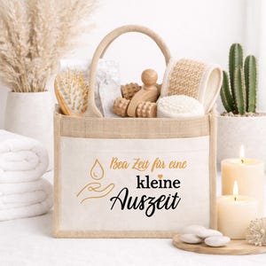Könnte beinhalten: Eine beigefarbene Tragetasche mit Spa-Produkten, darunter eine Holzbürste, Massage-Tools und ein Luffa. Die Tasche hat einen Griff und eine Vordertasche mit dem Text "Bea Zeit für eine kleine Auszeit" in Schwarz und Gold. Kerzen und Pflanzen sind im Hintergrund.