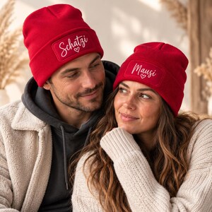 Könnte beinhalten: Zwei leuchtend rote Strickmützen mit rechteckigen Aufnähern auf der Vorderseite. Eine Mütze zeigt den weißen Schreibschrift-Text Schatzi mit einem kleinen Herzsymbol, die andere Mütze den weißen Schreibschrift-Text Mausi mit einem Herz.