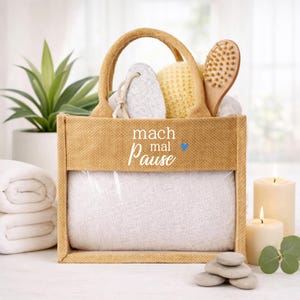 Könnte beinhalten: Eine beigefarbene Jute-Tragetasche gefüllt mit Spa-Utensilien, darunter ein weißes Handtuch, Bimsstein, Luffa und eine Holzbürste. Die Tasche hat eine transparente Front und den Schriftzug "mach mal Pause" in Weiß. Zwei brennende Kerzen und eine Pflanze sind im Hintergrund.