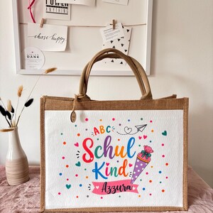May include: A rectangular jute tote bag with brown handles and a white front panel. The panel features colourful text reading ABC Schul Kind and the name Azzura, accompanied by a graphic of a cone, hearts, dots, and a paper airplane.