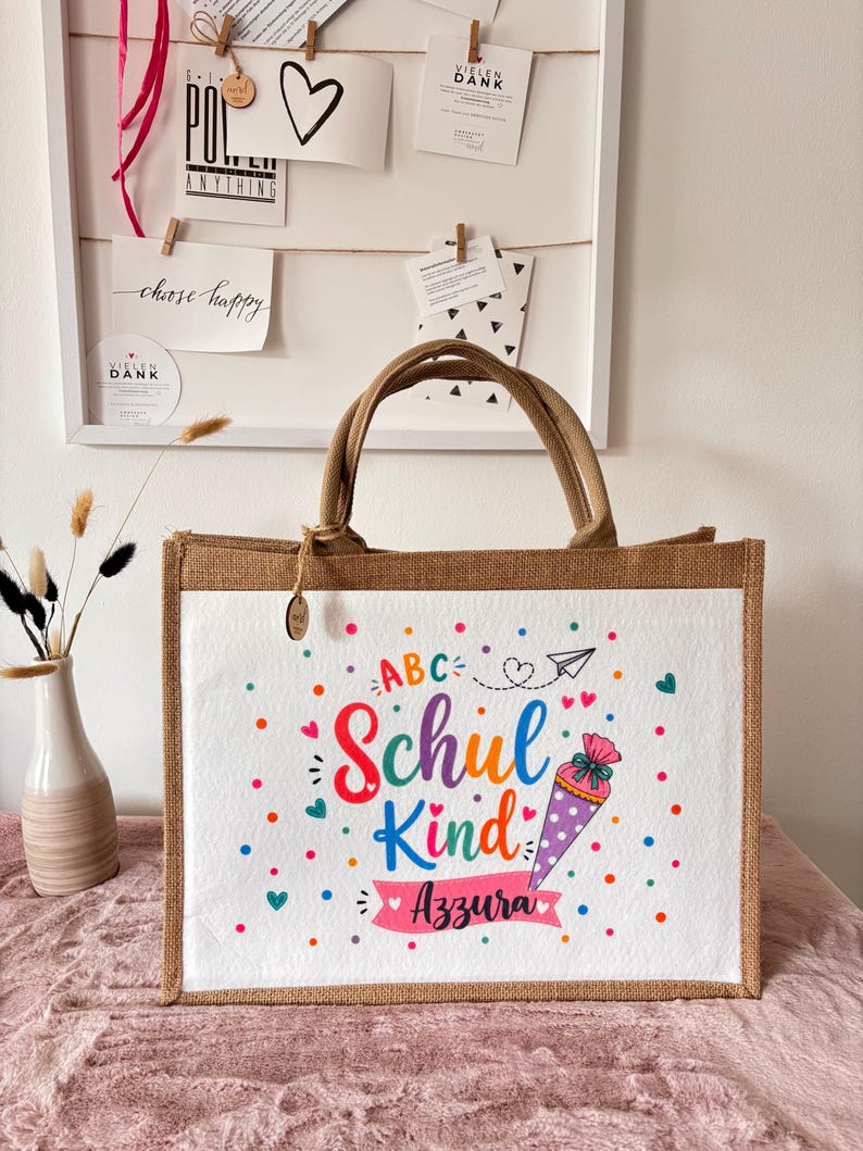 May include: A rectangular jute tote bag with brown handles and a white front panel. The panel features colourful text reading ABC Schul Kind and the name Azzura, accompanied by a graphic of a cone, hearts, dots, and a paper airplane.