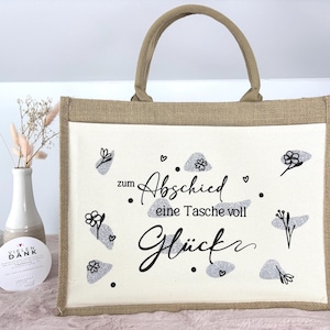 Pu&ograve; includere: Una borsa tote rettangolare con un telaio in juta beige e un pannello centrale bianco. Il pannello presenta il testo tedesco "zum Abschied eine Tasche voll Gl&uuml;ck" in scrittura nera, insieme a disegni floreali e a forma di nuvola grigia. La borsa ha un manico beige.