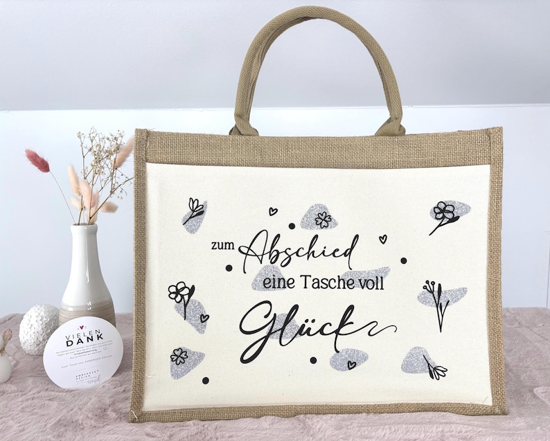 Pu&ograve; includere: Una borsa tote rettangolare con un telaio in juta beige e un pannello centrale bianco. Il pannello presenta il testo tedesco "zum Abschied eine Tasche voll Gl&uuml;ck" in scrittura nera, insieme a disegni floreali e a forma di nuvola grigia. La borsa ha un manico beige.