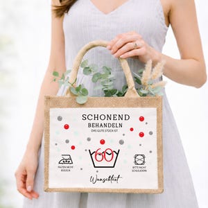 Puede incluir: Un bolso de yute rectangular con un panel frontal blanco que presenta el texto en alem&aacute;n SCHONEND BEHANDELN, DAS GUTE ST&Uuml;CK IST, FALTEN NICHT B&Uuml;GELN, BITTE NICHT SCHLEUDERN y WUNSCHTEXT, junto con un 60 rojo dentro de un s&iacute;mbolo de lavado y puntos.