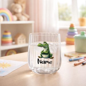 Könnte beinhalten: Ein klares, stieloses Glas, gefüllt mit Wasser, mit einem Cartoon-Alligator-Aufkleber. Der Alligator ist grün und weiß, mit dem Wort "Name" in schwarzer Schrift darunter. Das Glas hat vertikale Rillen.