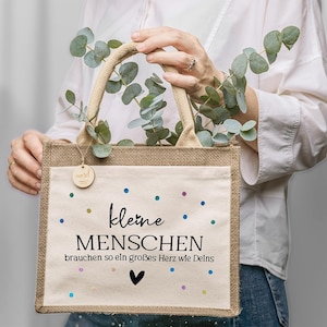 Könnte beinhalten: Eine rechteckige Tragetasche aus Jute mit einer cremefarbenen Frontplatte. Auf der Platte steht der deutsche Text "kleine MENSCHEN brauchen so ein großes Herz wie Deins" mit bunten Punkten und einem schwarzen Herz. Die Tasche hat einen Jute-Griff.
