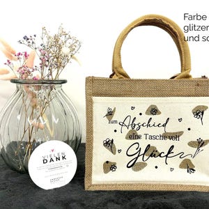 Pu&ograve; includere: Una piccola borsa tote rettangolare in iuta con un pannello frontale color crema con la scritta "zum Abschied eine Tasche voll Gl&uuml;ck" in nero e dettagli glitterati dorati. La borsa ha un manico in iuta abbinato.