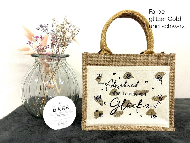 Pu&ograve; includere: Una piccola borsa tote rettangolare in iuta con un pannello frontale color crema con la scritta "zum Abschied eine Tasche voll Gl&uuml;ck" in nero e dettagli glitterati dorati. La borsa ha un manico in iuta abbinato.