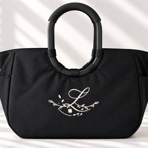 Könnte beinhalten: Eine schwarze Tragetasche mit einem starren, abgerundeten schwarzen Griff und Seitentaschen. Auf der Vorderseite der Tasche befindet sich ein weißes Grafikdesign, das aus einem kursiven Buchstaben L besteht, umgeben von botanischen Zweigen.
