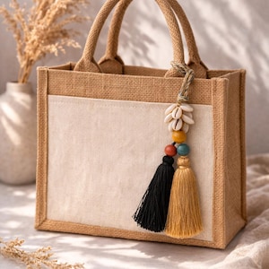 Könnte beinhalten: Kleine, rechteckige Tragetasche aus Jute mit einer cremefarbenen Vordertasche. Die Tasche hat zwei Griffe und ist mit einem dekorativen Quasten- und Perlenanhänger verziert. Der Anhänger enthält Muscheln, bunte Perlen und schwarze und goldene Quasten.