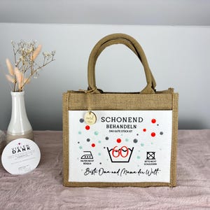 Puede incluir: Un bolso de yute marr&oacute;n con un panel frontal blanco que presenta el texto SCHONEND BEHANDELN DAS GUTE ST&Uuml;CK IST, un s&iacute;mbolo de lavado con el n&uacute;mero 60 y el texto Beste Oma und Mama der Welt, junto con acentos circulares rojos y grises.