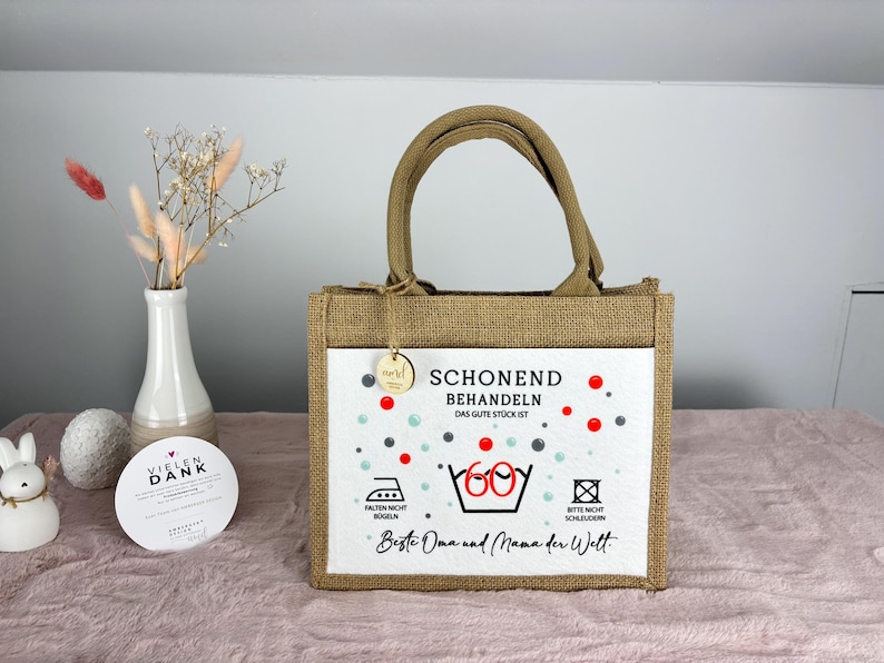 Puede incluir: Un bolso de yute marr&oacute;n con un panel frontal blanco que presenta el texto SCHONEND BEHANDELN DAS GUTE ST&Uuml;CK IST, un s&iacute;mbolo de lavado con el n&uacute;mero 60 y el texto Beste Oma und Mama der Welt, junto con acentos circulares rojos y grises.