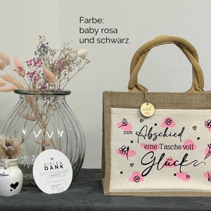 Pu&ograve; includere: Una borsa tote in iuta beige con un pannello frontale bianco con disegni floreali rosa e le parole "zum Abschied eine Tasche voll Gl&uuml;ck". La borsa ha un manico intrecciato e una piccola etichetta di legno. Un vaso con fiori secchi &egrave; a sinistra.