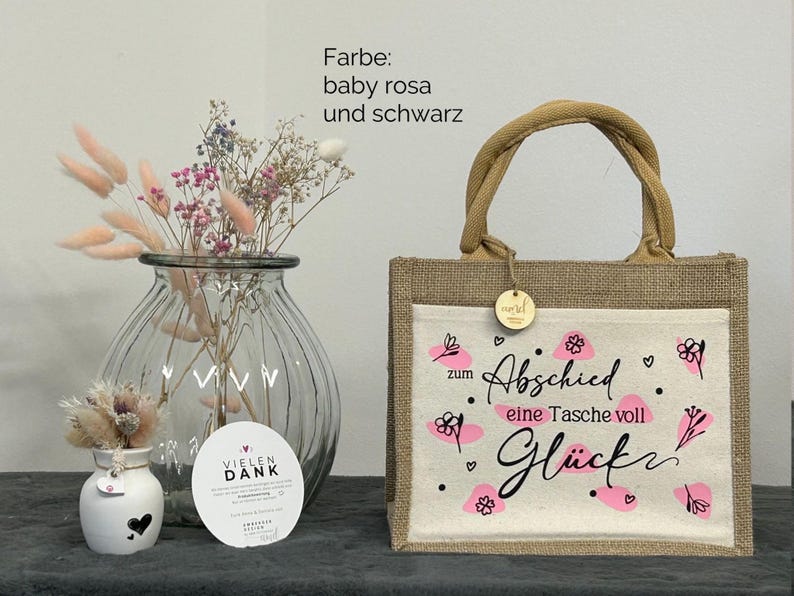 Pu&ograve; includere: Una borsa tote in iuta beige con un pannello frontale bianco con disegni floreali rosa e le parole "zum Abschied eine Tasche voll Gl&uuml;ck". La borsa ha un manico intrecciato e una piccola etichetta di legno. Un vaso con fiori secchi &egrave; a sinistra.
