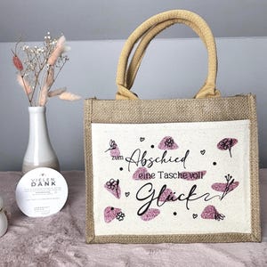 Pu&ograve; includere: Una borsa tote in iuta con un pannello frontale color crema con il testo tedesco "zum Abschied eine Tasche voll Gl&uuml;ck" in scrittura nera e accenti glitter rosa. La borsa ha manici color cuoio e un corpo in iuta naturale.