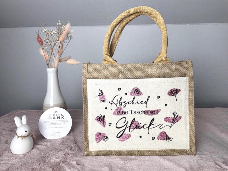 Pu&ograve; includere: Una borsa tote in iuta con un pannello frontale color crema con il testo tedesco "zum Abschied eine Tasche voll Gl&uuml;ck" in scrittura nera e accenti glitter rosa. La borsa ha manici color cuoio e un corpo in iuta naturale.