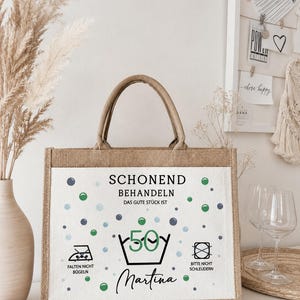 Puede incluir: Un bolso tote rectangular de yute con un panel frontal blanco que presenta el texto "SCHONEND BEHANDELN DAS GUTE ST&Uuml;CK IST 50" y "Martina" en negro, junto con iconos de cuidado de lavado y puntos azules y verdes sobre una base de tela beige.