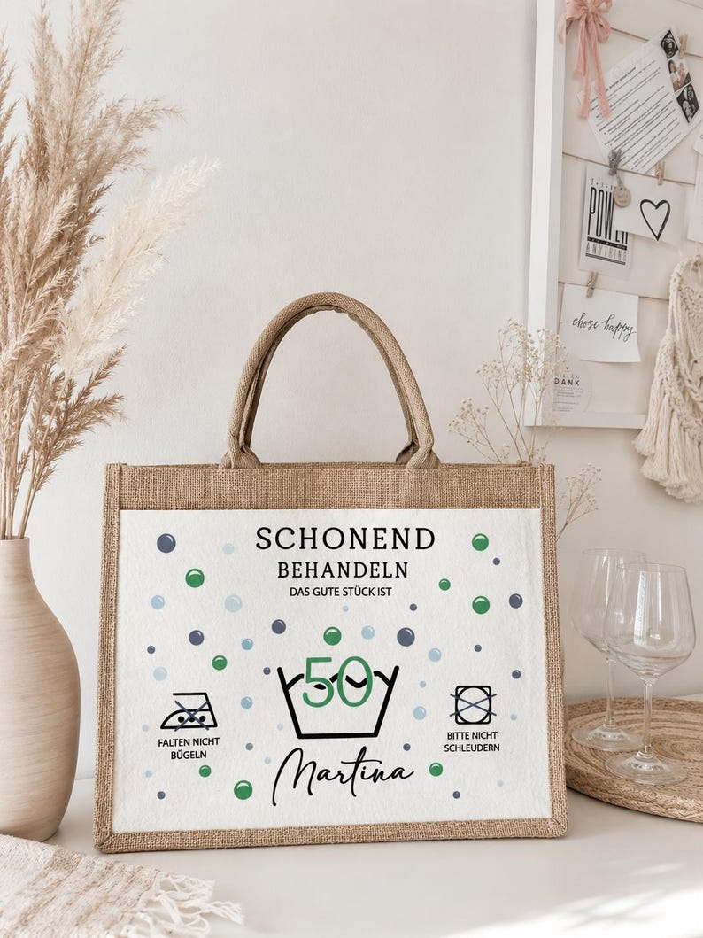Puede incluir: Un bolso tote rectangular de yute con un panel frontal blanco que presenta el texto "SCHONEND BEHANDELN DAS GUTE ST&Uuml;CK IST 50" y "Martina" en negro, junto con iconos de cuidado de lavado y puntos azules y verdes sobre una base de tela beige.