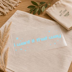 Könnte beinhalten: Ein rechteckiger, transparenter Vinyl-Aufkleber mit dem hellblauen Schriftzug i want it that way, umgeben von kleinen Sternsymbolen. Der Aufkleber liegt auf einem hellbeigen Stoff neben einem kleinen Beutel mit der Aufschrift DIY MIT LIEBE.