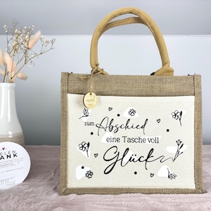 Pu&ograve; includere: Una borsa tote in iuta con un pannello frontale color crema con il testo tedesco "zum Abschied eine Tasche voll Gl&uuml;ck" in scrittura nera, che significa "una borsa piena di fortuna". Ha un manico in iuta e una piccola etichetta di legno.